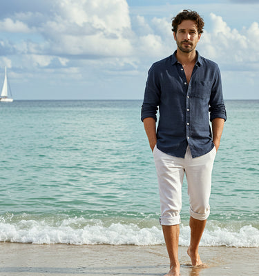 A man standing barefoot on the beach wearing a navy blue button down linen shirt paired with white linen pants that are rolled up below the knees.