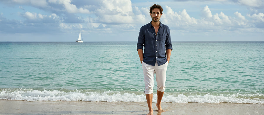 A man standing barefoot on the beach wearing a navy blue button down linen shirt paired with white linen pants that are rolled up below the knees.