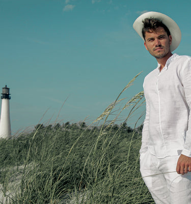 A man wearing an all white 100% linen outfit standing along a sunny coastline with a lighthouse and a mountainous village in the background.