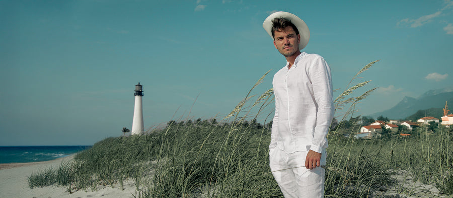 A man wearing an all white 100% linen outfit standing along a sunny coastline with a lighthouse and a mountainous village in the background.