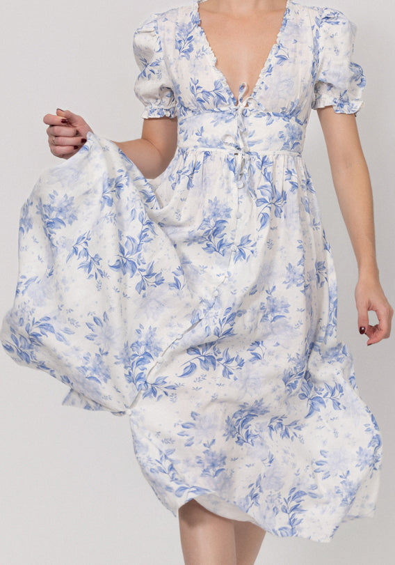 Woman wearing a linen white floral dress holding a white bag with blue floral pattern on a plain background